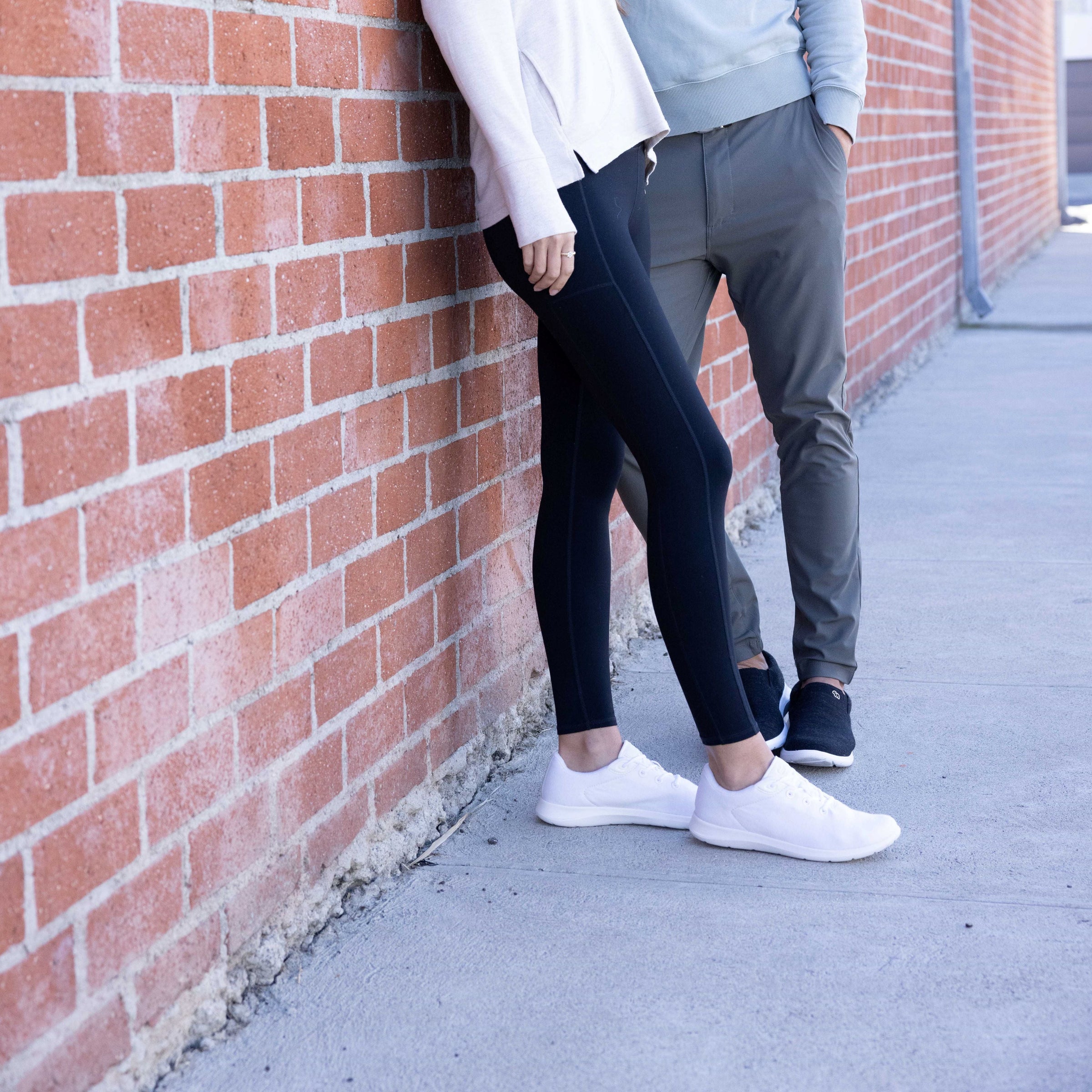 Two people standing against a brick wall, one is wearing white lace-ups and the other is wearing black slip-ons with white soles.